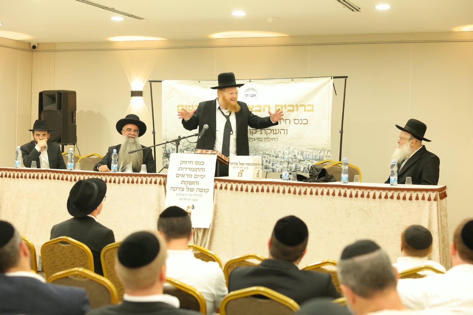 הרב מרון שליט״א נושא דברי חיזוק מאחורי הדוכן, שולחן הכבוד והקהל באולם